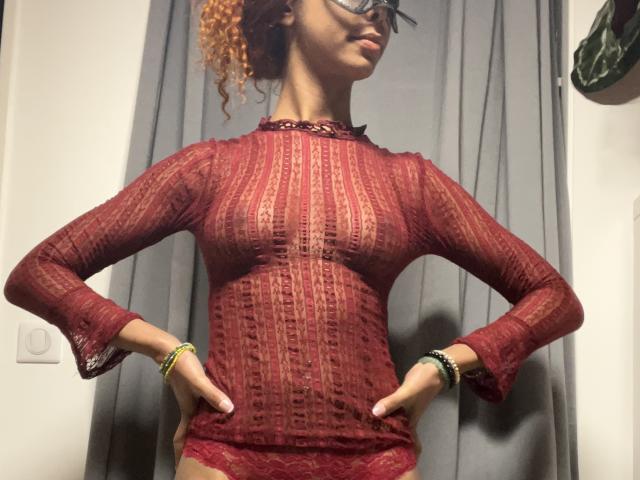 KaylaSun with curly red hair, glasses, wearing a red lace top, hands on hips, showing her tits, gray curtains in the background, green and black bracelets on her wrists.