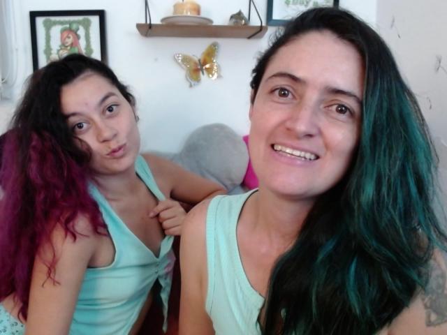 LunaVenus, con cabello rosa y negro, frunce los labios y lleva puesto un top verde, junto a LunaVenus, con cabello verde y negro, que sonríe. Ambas tienen senos pequeños y están sin sujetador..