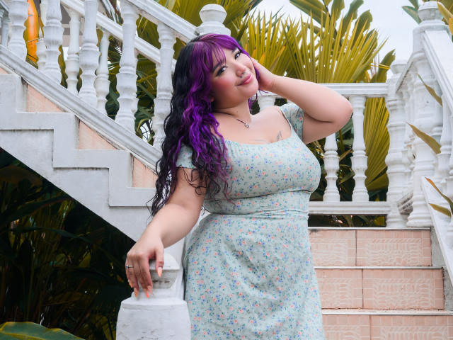 HonnyBlaice, com cabelo roxo e preto, vestindo um vestido floral, em pé ao lado de escadas brancas, com a mão na cabeça, olhando para a câmera, exibindo seus seios grandes e traseiro curvilíneo..