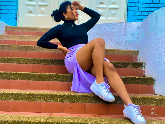 SophieSonyas with dark skin, black turtleneck, purple skirt, white sneakers, and curly hair, sitting on red brick stairs, hand on head, legs crossed, showing her ass.