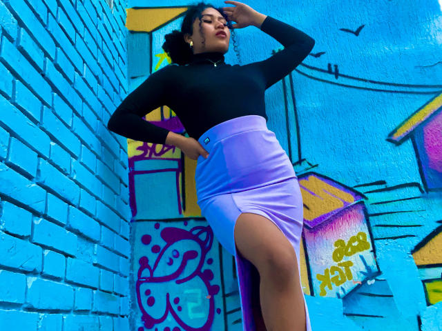 SophieSonyas with curly hair, black long-sleeve top, and purple skirt with a thigh-high slit, standing against a bright blue brick wall, hand on hip, looking down.