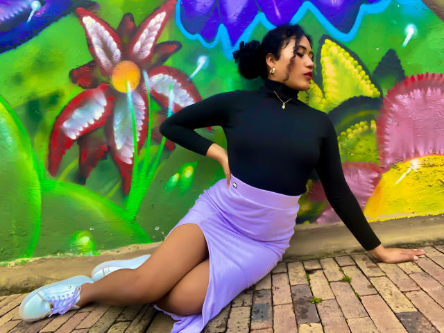 SophieSonyas, com pele marrom média, blusa preta, saia roxa, tênis brancos e cabelo preto cacheado, sentada em calçada de tijolos, com a mão na cintura, exibindo suas nádegas e coxas..