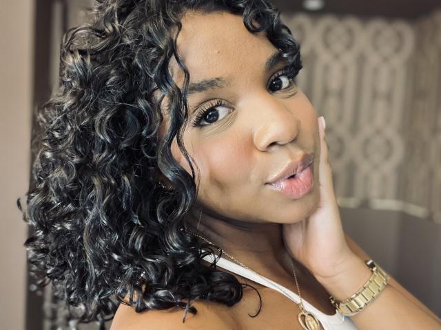 LisaMendez with curly black hair, brown skin, and pouty pink lips, touching her cheek with a hand, wearing a white top and gold watch, looking at the camera.
