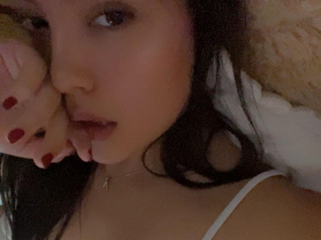 IssaPuasini with pouty lips, holding a plush toy to her face, wearing a white spaghetti strap top, dark hair, red nails, soft brown skin, small necklace.
