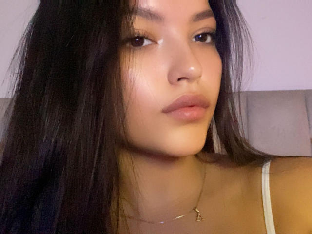 IssaPuasini has glossy, dark brown hair, full lips, and brown eyes, wearing a white tank top with a gold necklace. Her skin is smooth and slightly shiny.