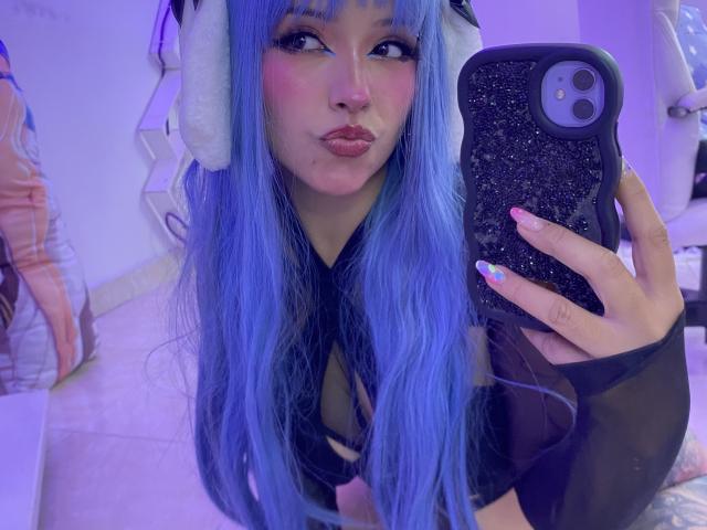 CarlaMiceli with blue hair, pouty lips, wearing a black mesh top, holding a glittery phone, wearing white furry headphones, in a purple-lit room, showing off colorful nails.