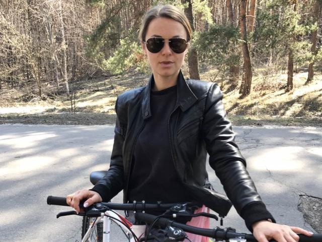 BelleLisaG with sunglasses, black leather jacket, black top, pink skirt, riding a bicycle, hands on bars, forest background, day, serious expression, brown hair in a ponytail.