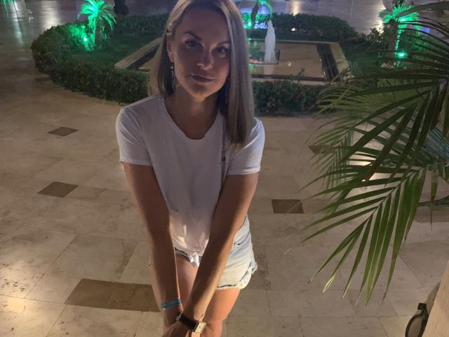 BelleLisaG with blonde hair, white tee, and denim shorts, standing, hands on knees, smiling, green plant in background, evening light, marble floor, wrist bracelet, slightly visible tits.