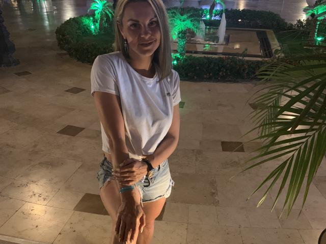 BelleLisaG, avec des cheveux blonds, un t-shirt blanc et un short en jean, debout, les bras croisés, souriante, les mains sur les genoux, jardin en arrière-plan éclairé en vert, feuilles de palmier, nuit, sol en marbre, décontractée, attrayante..