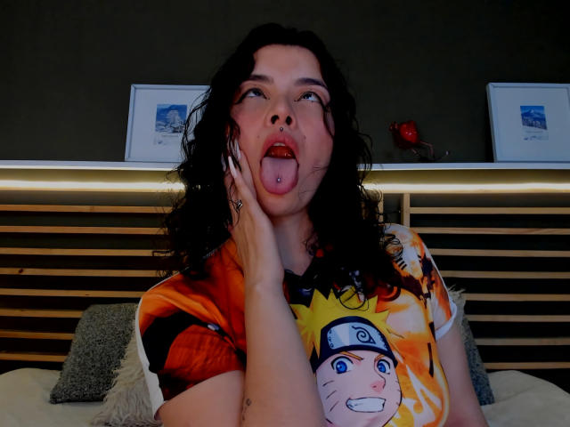 LilibethHot69 con capelli neri ricci, la lingua fuori, piercing rosa sulla lingua, indossa una maglietta di Naruto, una mano sul viso, guarda in alto, a letto con una testiera in legno, cuscini grigi..