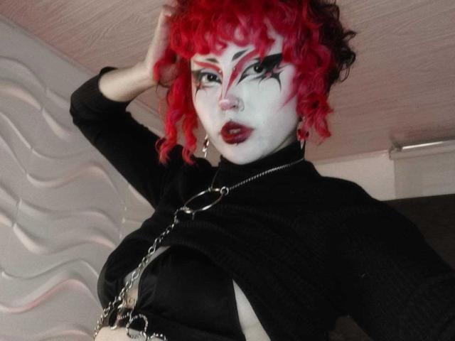 GothVioleth con capelli ricci rossi vivaci, viso truccato di bianco, eyeliner nero e rossetto, indossa una maglia nera, una catena e un anello, in posa con la mano tra i capelli. Seni visibili..