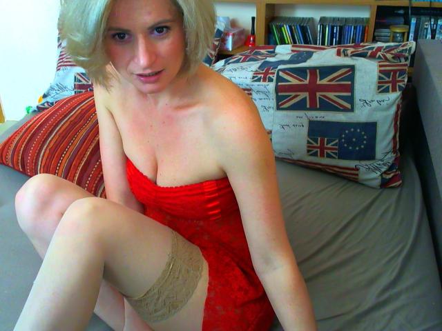 ChatePoilue with blonde hair, wearing a red lace dress, exposing her tits, and beige thigh-highs, sitting on a bed with Union Jack pillows.