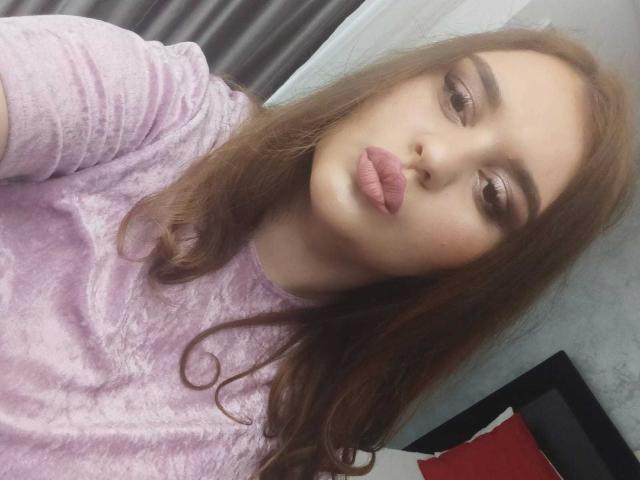SarahClimber with pouty pink lips, brown eyes, and wavy brown hair, wearing a pink velvet shirt, looking at the camera. Boobs are not visible.