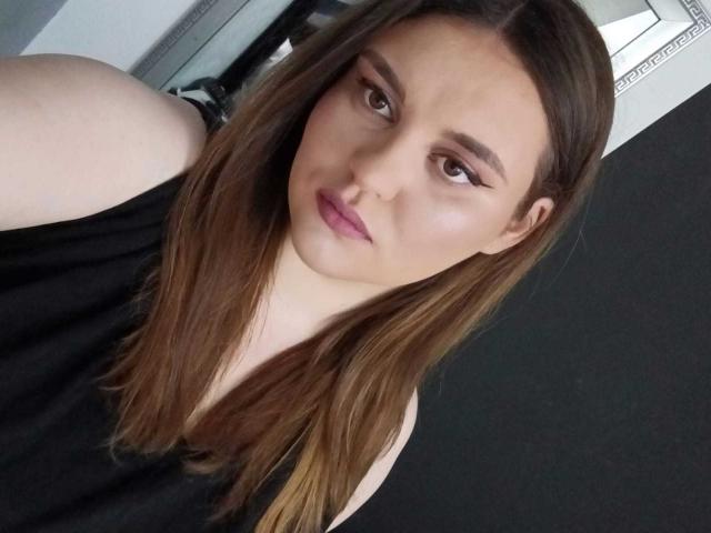 SarahClimber has brown hair, a pale skin tone, and is wearing a black sleeveless top. She has pink lipstick and eyeliner, looking at the camera with a slight pout.