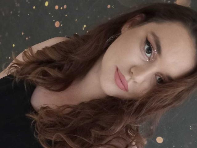 SarahClimber has wavy brown hair, blue eyes, and pink lipstick. She's wearing a black top, with a subtle, confident expression, and small hoop earrings.