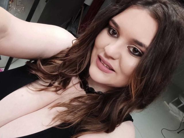 SarahClimber with wavy brown hair, wearing a black top, showing large tits, smiling with pink lipstick, brown eyes, and a black bead necklace, taking a selfie.
