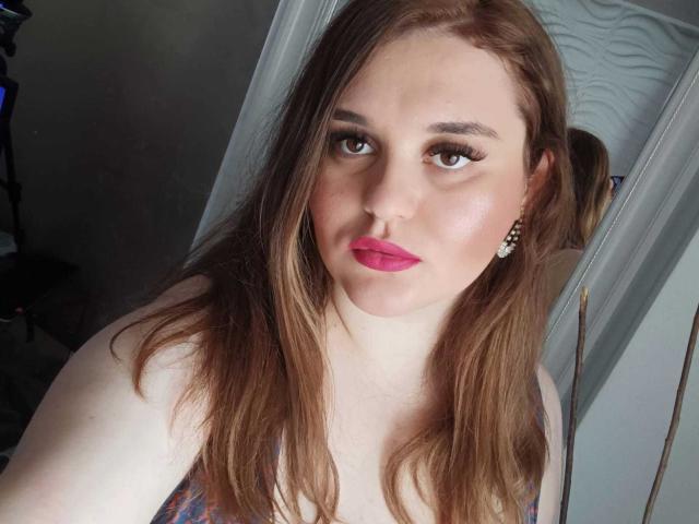SarahClimber has pale skin, long brown hair, pink lipstick, and wears a patterned top. She has eyeliner and pearl earrings, looking directly at the camera with a slight smile.