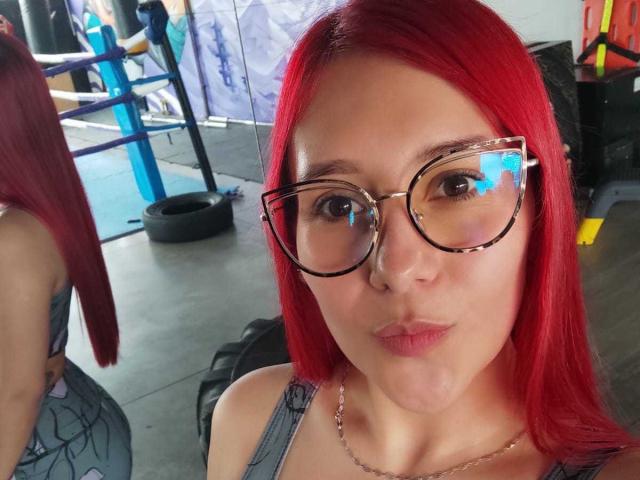 JuliaRouse with red hair, pouty lips, and tortoiseshell glasses, wearing a gray tank top, in a gym. Her tits are small, and she's pouting at the camera.