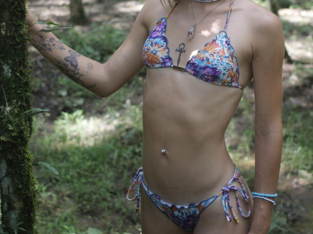 BloomSun indossa un bikini floreale colorato, mostrando una pelle abbronzata e snella, con tatuaggi sul braccio e un piercing all'ombelico. È in piedi in una foresta, toccando un albero ricoperto di muschio..