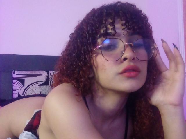 AmaiaEscotet with curly brown hair, glasses, red lipstick, and black nails, laying on her stomach, showing her tits, looking at the camera, hand near her face.