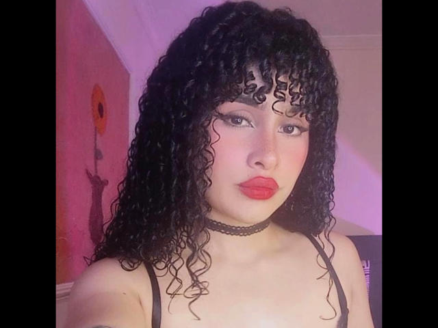 Amaia Escotet, con cabello negro rizado, lápiz labial rojo, collar negro y sujetador negro, mirando a la cámara, mostrando sus pechos, iluminación rosada, expresión seria, plano medio..