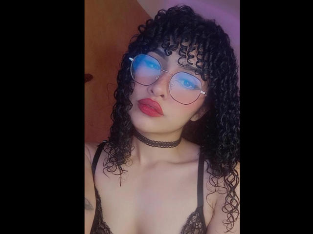 Amaia Escotet mit lockigem, schwarzem Haar, rotem Lippenstift, blau getönten Brillen, einer schwarzen Halskette und einem Spitzenoberteil, wobei kleine Brüste zu sehen sind, vor einem dunklen Hintergrund..