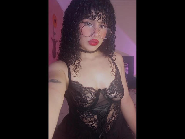 Amaia Escotet mit lockigem, schwarzem Haar, roten Lippen und Brille, trägt ein schwarzes Spitzendessous-Kleid, das ihre Brüste, eine Halskette und ein Tattoo an ihrem linken Arm zeigt..