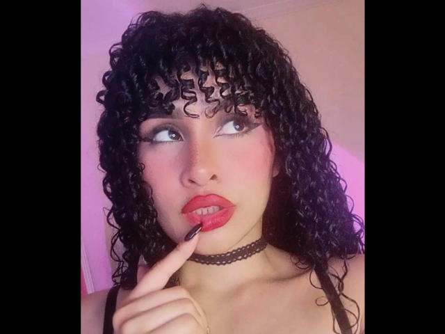 AmaiaEscotet with curly black hair, red lipstick, black choker, and finger touching her lip, looking up with blue eyes, showing her small tits and cute face.