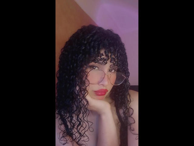Amaia Escotet mit lockigem, schwarzem Haar, die runde Brillen und roten Lippenstift trägt, lehnt ihren Kopf in ihre Hand und zeigt ihre nackten Schultern, mit einem sexy und selbstbewussten Ausdruck..