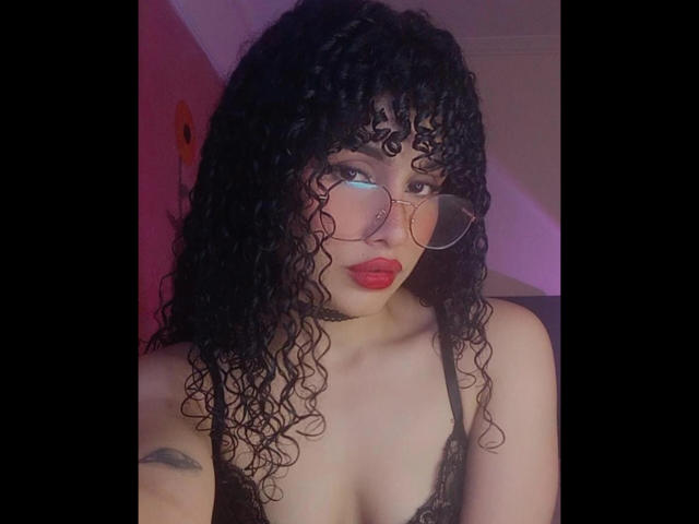 Amaia Escotet, com cabelo preto encaracolado, batom vermelho e óculos, vestindo um sutiã de renda preto, mostrando seus pequenos seios, olhando para a câmera com um leve sorriso..