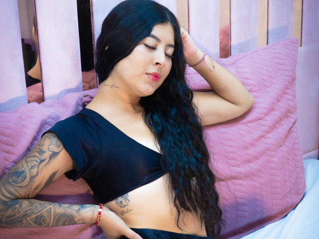 NicoleCollins with long black hair, black top, tattoos on arm and side, red lipstick, eyes closed, pink quilted pillow, wooden bed, right arm behind head, left arm on pillow.