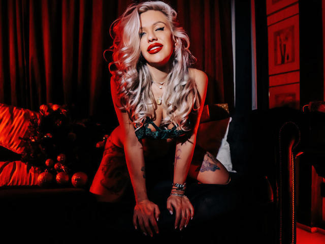 AnastasiaDeepp with platinum blonde curls, red lipstick, green lace bra, tattoos, and black pants, kneeling on a dark couch, hands on seat, red and black lighting, smiling seductively.