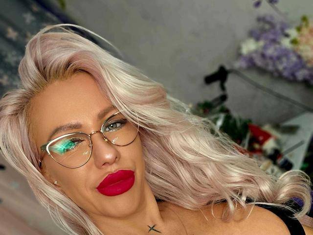 AnastasiaDeepp with platinum blonde hair, red lipstick, green-tinted glasses, and a black top, showing a cross tattoo on her neck. Her green eyes are slightly squinted.