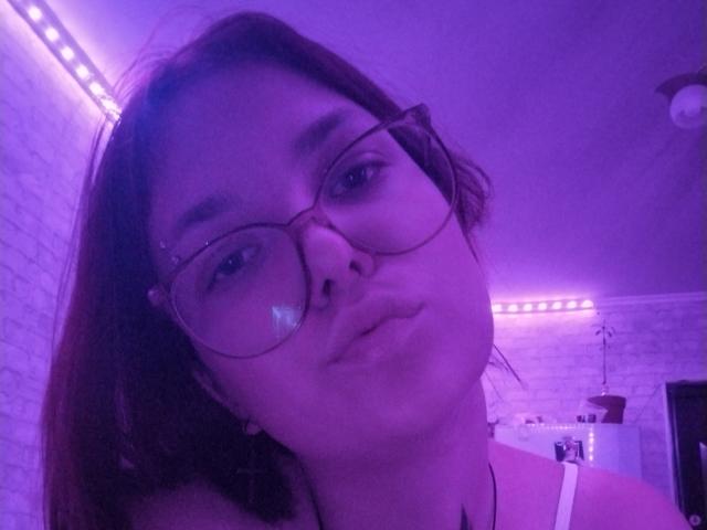Gene Kim mit Brille, leicht geschürzten Lippen und einem leichten Grinsen, unter violettem Licht, zeigt ihr Gesicht und einen Hauch ihres Halses mit einer schwarzen Halskette..