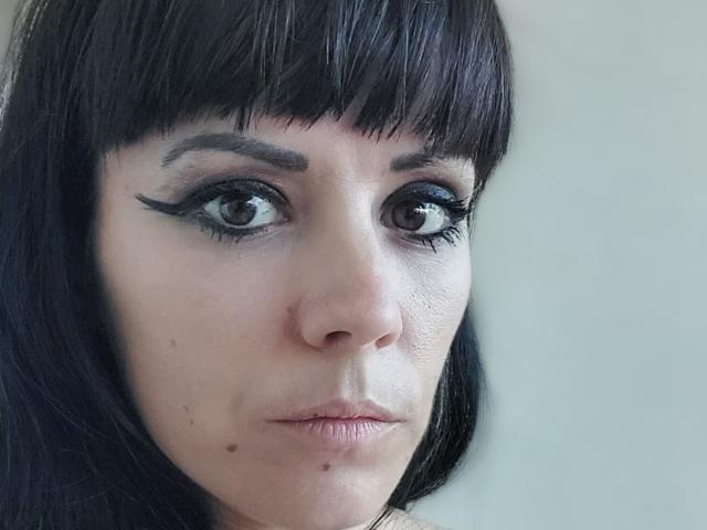OrianaLaFrancaise a un trait d'eye-liner noir foncé, une petite tâche sous son œil droit, et une frange noire droite qui recouvre son front. Son expression est sérieuse, avec les lèvres légèrement entrouvertes..