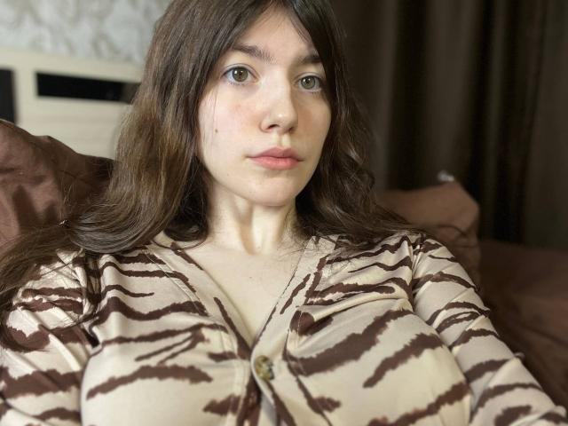 VinnaSkyHott, met lang bruin haar, groene ogen en lichte huid, draagt een gebreide vest met zebravlekken, waarbij haar borsten zichtbaar zijn, en zit op een bruin bed, kijkend in de camera..