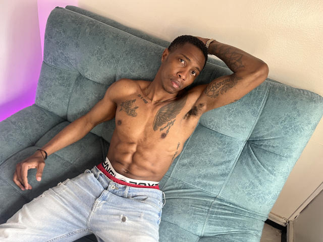 Daviicii ohne Hemd, mit Tattoos, in blauen Jeans und einem PUMA-Hüftgurt, liegt auf einem grünen Sofa, der rechte Arm ist hinter dem Kopf, der linke Arm ist nach unten gestreckt, und er schaut in die Kamera..