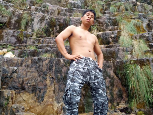 MarcoVernicos, sin camisa, de pie con las manos en las caderas, usando pantalones de camuflaje, mirando hacia arriba, una escalera de piedra con césped verde al fondo..