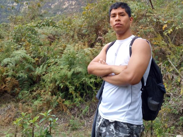 MarcoVernicos, con los brazos cruzados, vistiendo una camiseta blanca sin mangas, una mochila negra y pantalones de camuflaje, está de pie en un área verde y frondosa con helechos. Tiene el pelo corto y negro y una expresión seria..