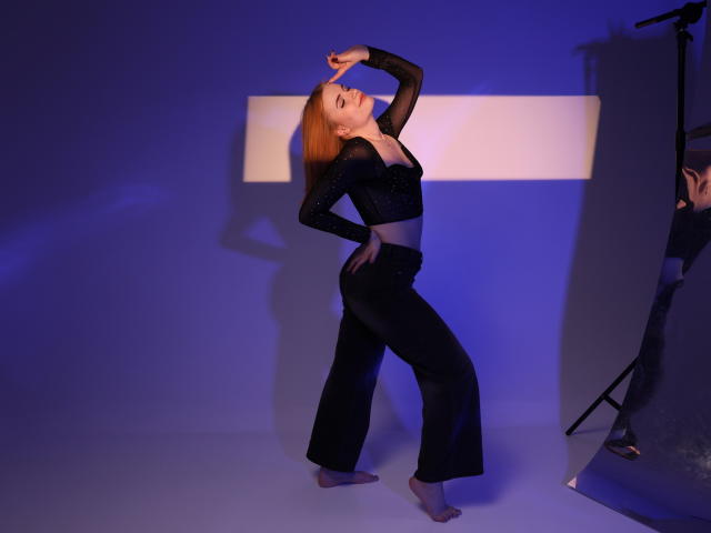 MargaritaKis with red hair, black sheer top, and black pants, barefoot, standing on one leg, arm raised, blue and white lighting, photographer's stand on the right.