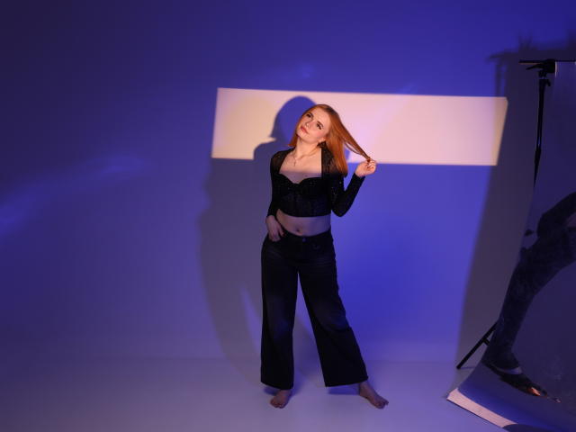 MargaritaKis with long red hair, holding a lock, wearing black lace bra and black pants, standing in blue light, shadow on wall, photographer's leg on right.