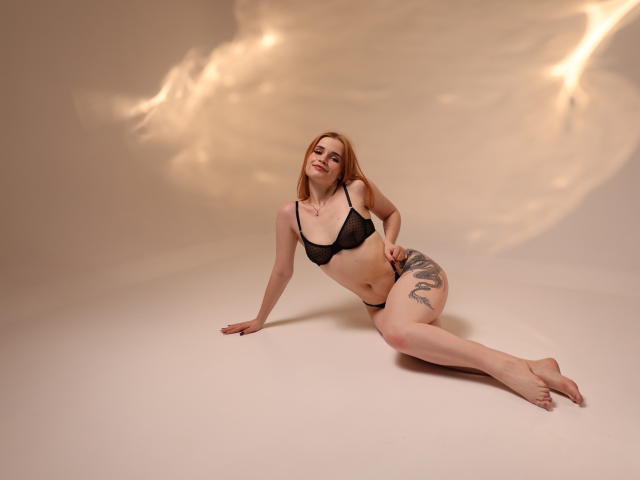 MargaritaKis with pale skin, red hair, black lace bra, black panties, tattoo on right thigh, sitting on white floor, leaning on right hand, legs extended, looking up.