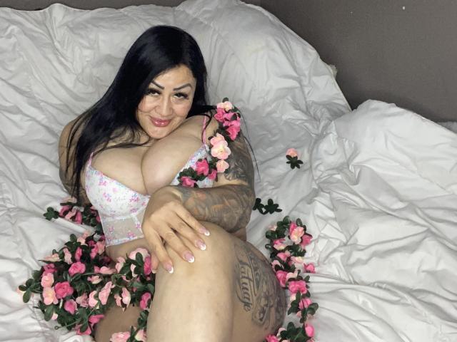 KittyHotLady69 con grandi seni, capelli neri, tatuaggi, reggiseno floreale rosa e fiori, sdraiata su un letto bianco, sorridente, gambe incrociate, tatuaggio sulla coscia, petali floreali rosa sparsi..