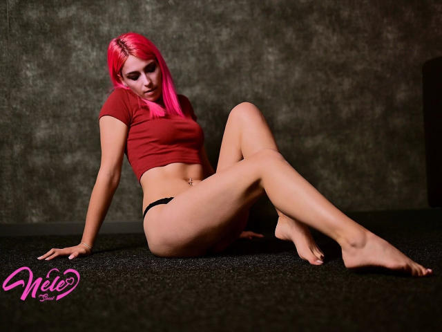 NeleSweet-hot with pink hair, red crop top, black panties, sitting on dark carpet, legs crossed, arms behind, looking down, showing her ass and a belly button piercing.