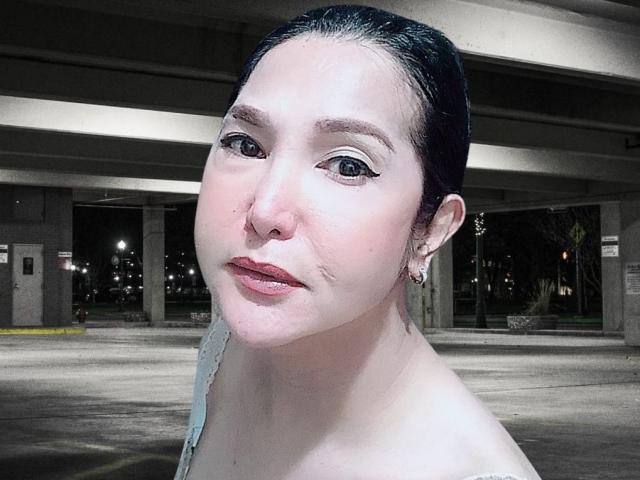 MistressKat with pale skin, dark hair in a bun, subtle eyeliner, pink lips, small nose, and small earrings, standing under a concrete overpass at night.
