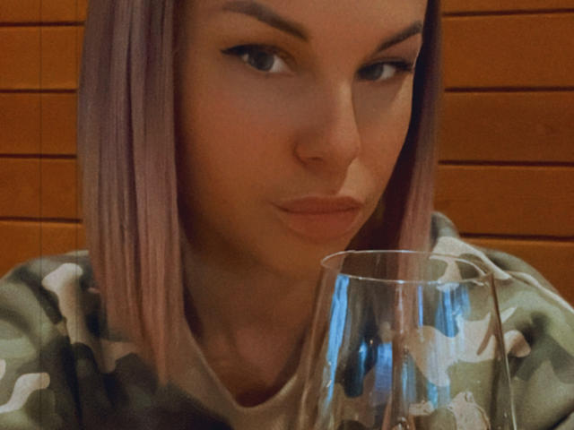 BelleLisaG with pink hair, green camo shirt, holding a glass, puckering lips, looking at camera, slight smile, brown wood background, eyeliner, soft lighting.