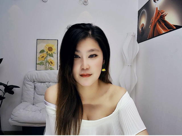 AilsaJade has long black hair, fair skin, and red lipstick, wearing a white off-shoulder top, yellow earrings, and a slight smile, showing her small tits.