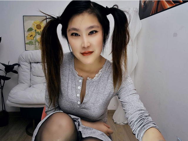 AilsaJade with pigtails, wearing a gray long-sleeve dress, kneeling, showing her tits, sitting on a white chair, smiling, red lipstick, white walls, wooden floor.