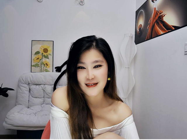 AilsaJade with long black hair, off-shoulder white top, smiling, showing her tits, in a room with a white chair and sunflower painting. Her ass is slightly visible.