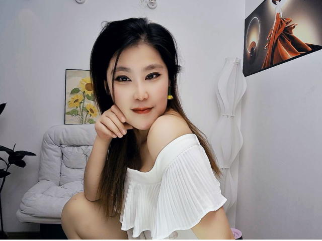 AilsaJade with dark hair, yellow earrings, white off-shoulder top, sitting, hand on chin, smiling, small tits, cute, Asian, indoors, white wall, chair, plant, artwork.
